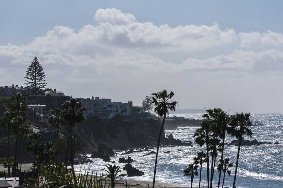 Rocky Southern California coastline with palm trees and buildings along the bluff under a partly cloudy sky